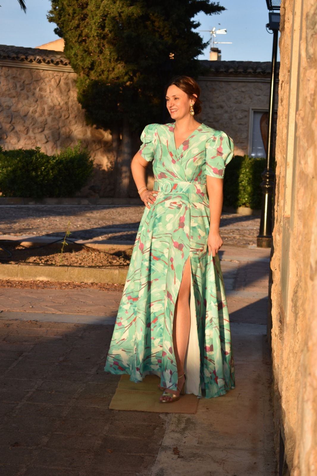 Mujer con un vestido verde estampado y una abertura alta, de pie afuera, con la mano en la cadera.