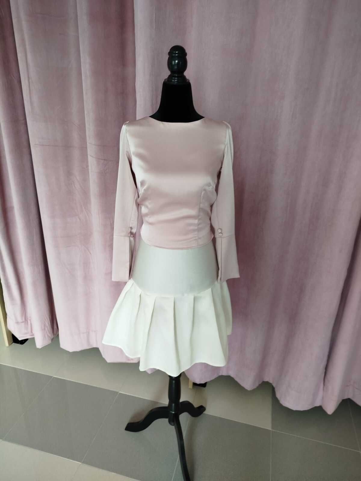 Un vestido sobre un maniquí negro. Top rosa pálido, falda plisada blanca, sobre cortinas rosas.