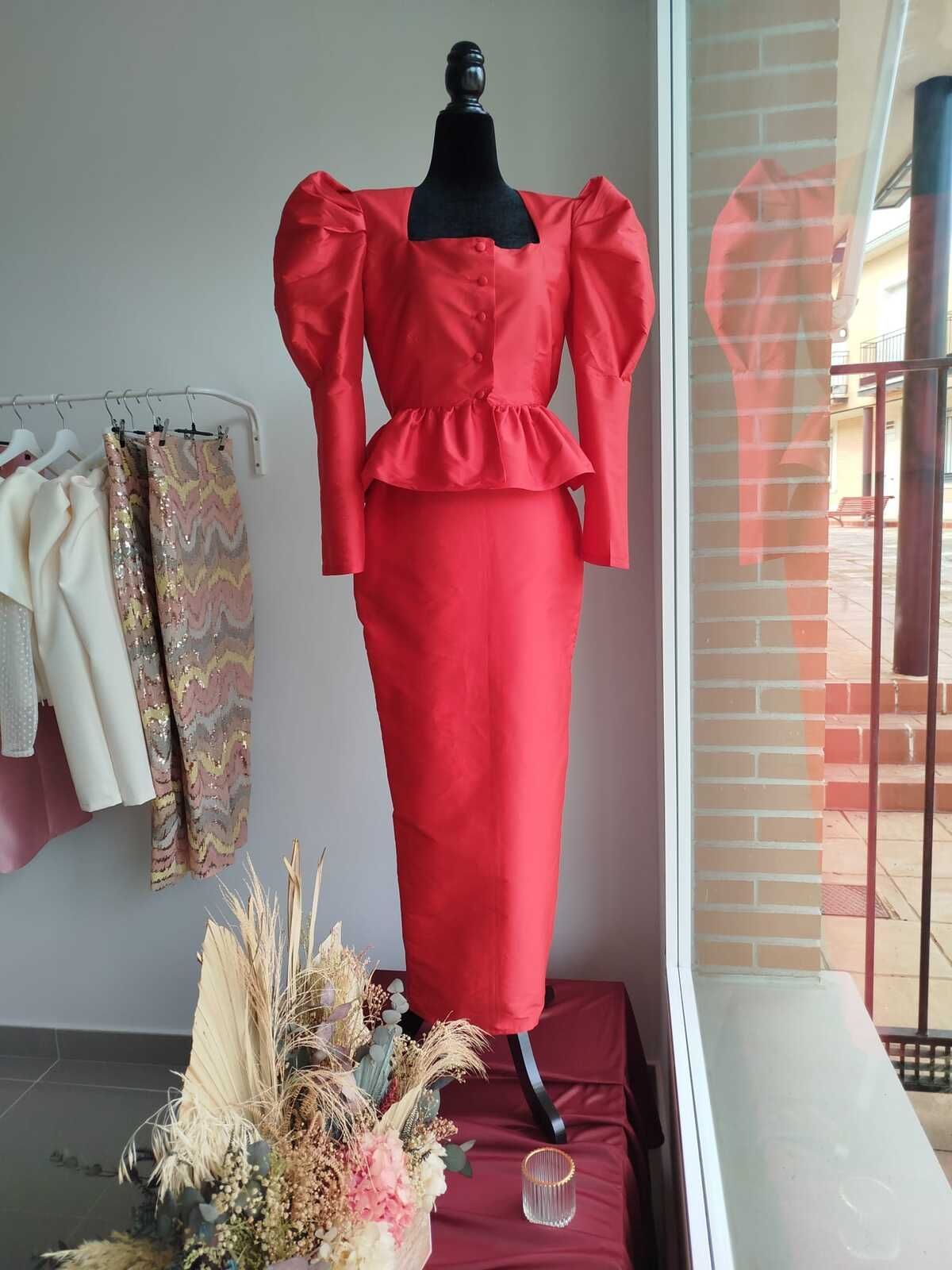 Vestido rojo sobre un maniquí. Tiene mangas abullonadas, peplum y falda larga. Interior, junto a una ventana.