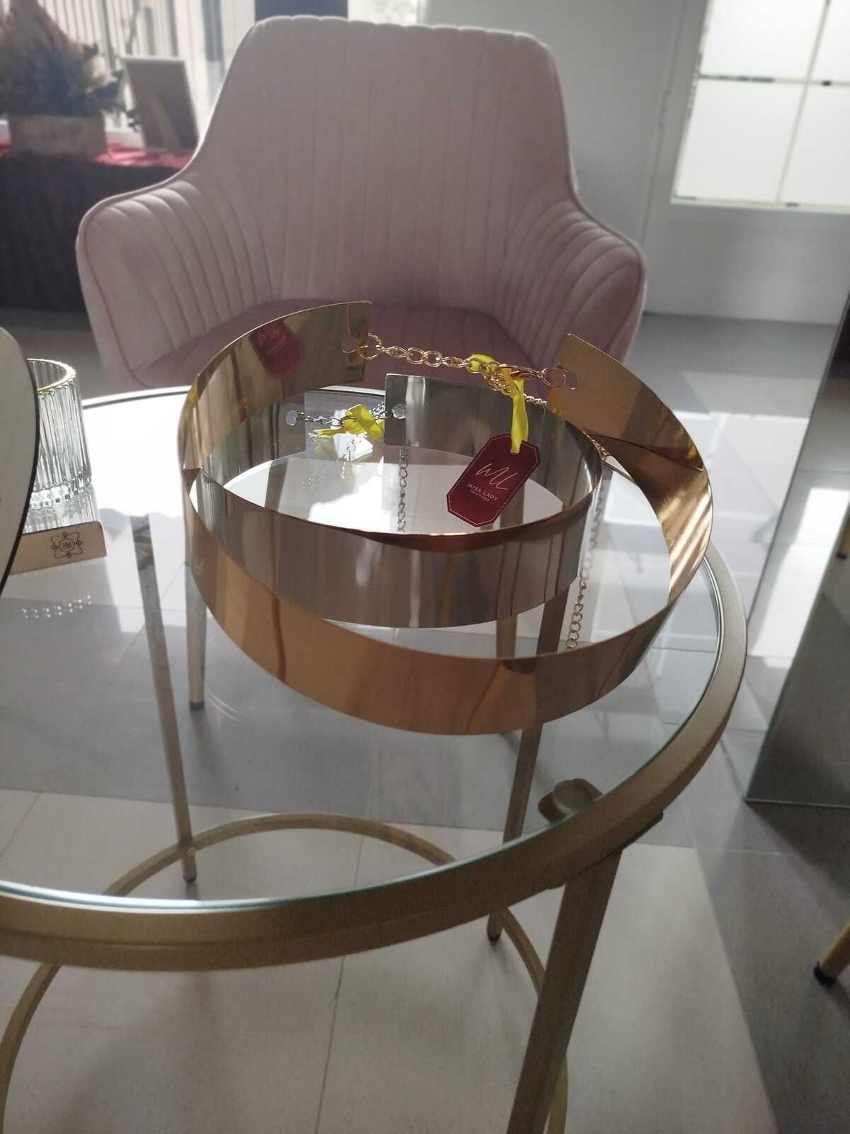 Una mesa redonda de cristal con marco dorado y anillos metálicos decorativos, un sillón rosa al fondo.