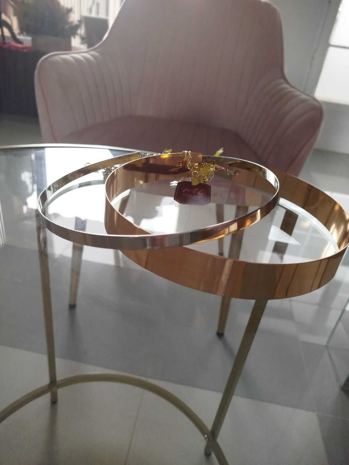 Mesa auxiliar dorada con tapa de cristal; un sillón rosa en el fondo.