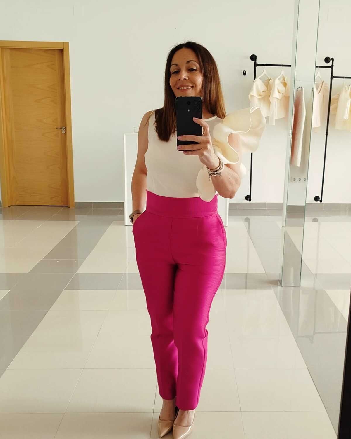 Mujer con top blanco y pantalones rosas, sosteniendo un teléfono frente a un espejo.