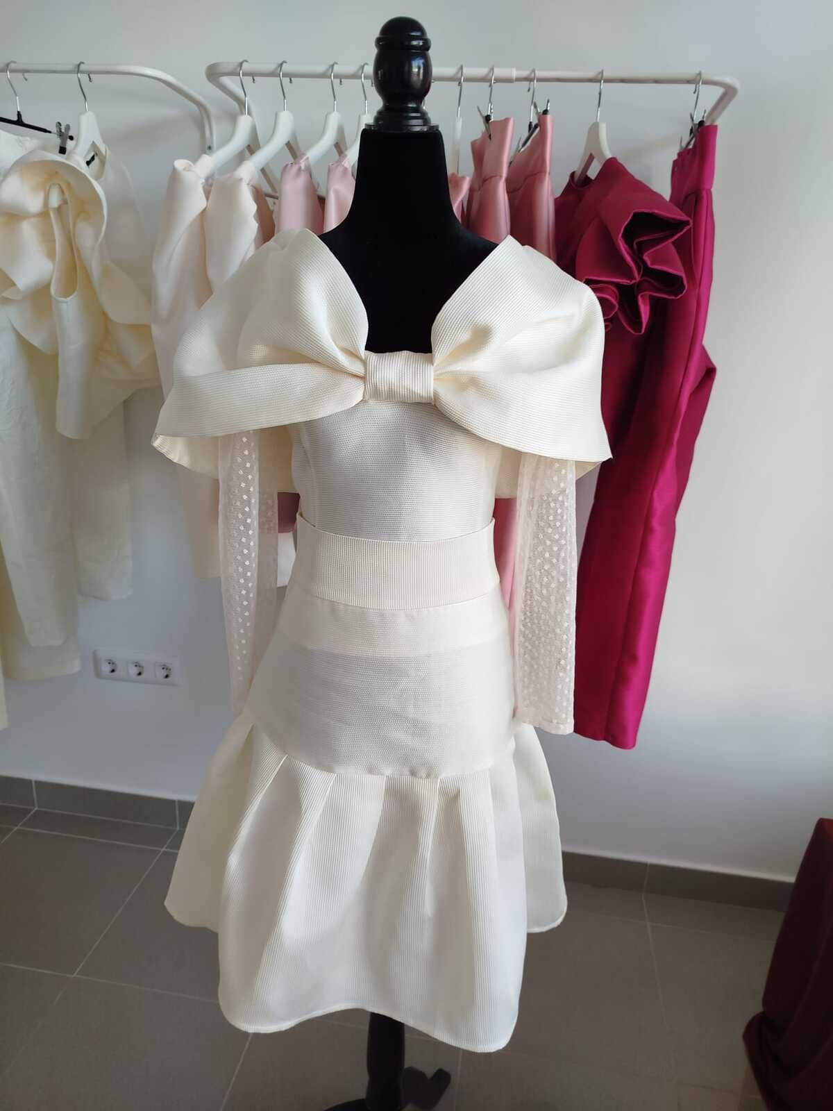 Vestido color crema sobre maniquí con escote de lazo grande, mangas largas y falda acampanada; perchero de vestidos al fondo.