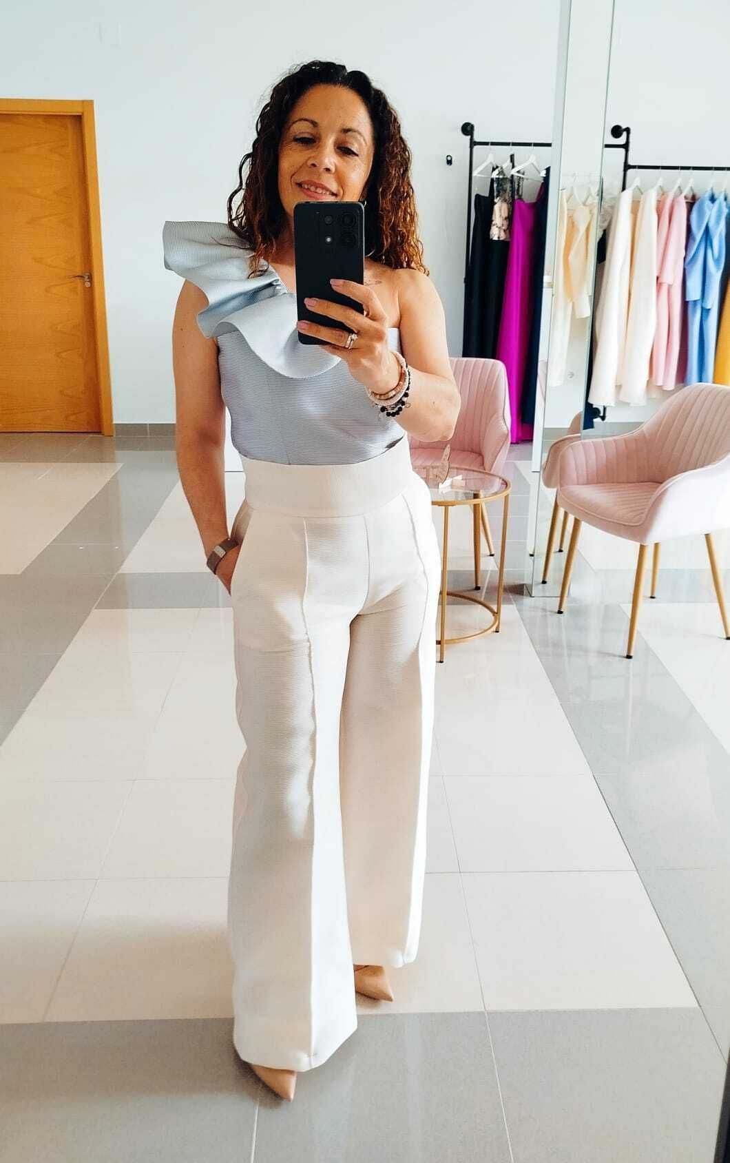 Una mujer con un top de un solo hombro y pantalones anchos se toma una selfie frente al espejo en un interior.