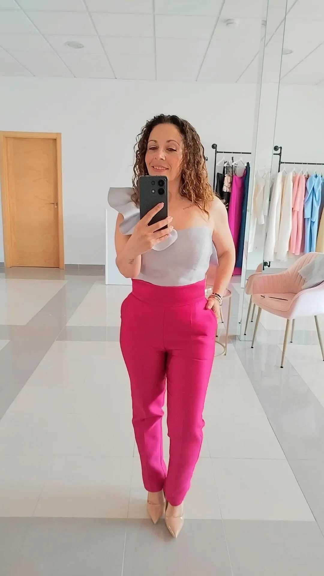 Mujer con top gris y pantalones rosas tomándose una selfie frente al espejo en una habitación luminosa.