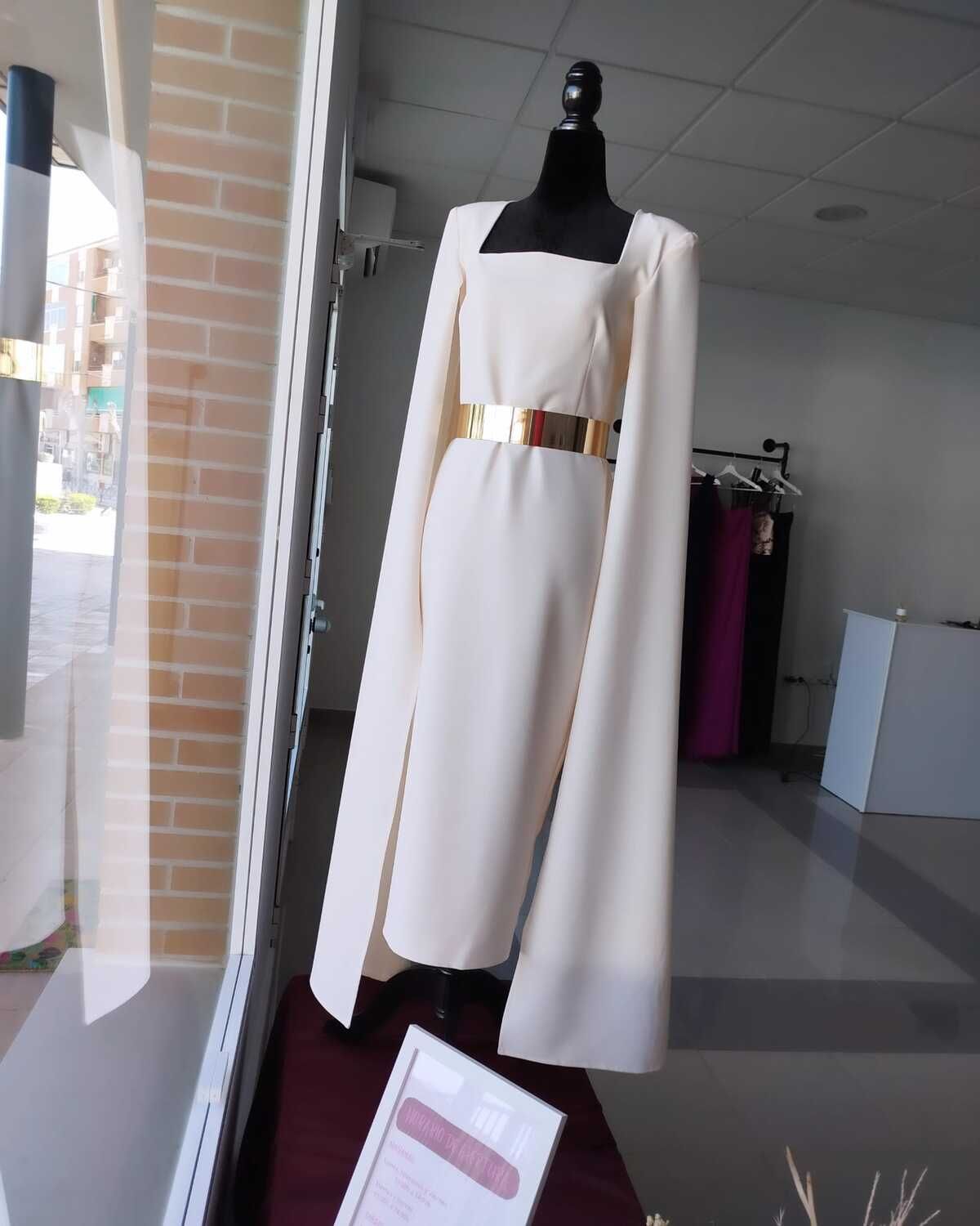 Vestido color marfil con cinturón dorado, escote cuadrado, mangas largas, sobre maniquí en escaparate de tienda.