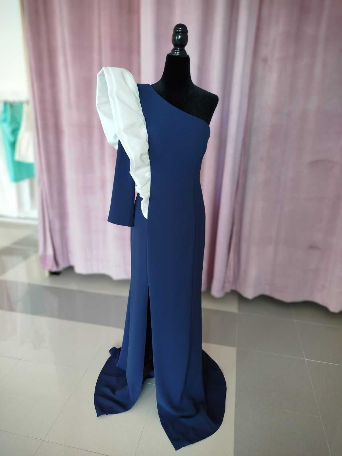 Vestido azul marino de un solo hombro con mangas blancas abullonadas sobre un maniquí. Pierna abierta y fondo de cortina rosa.