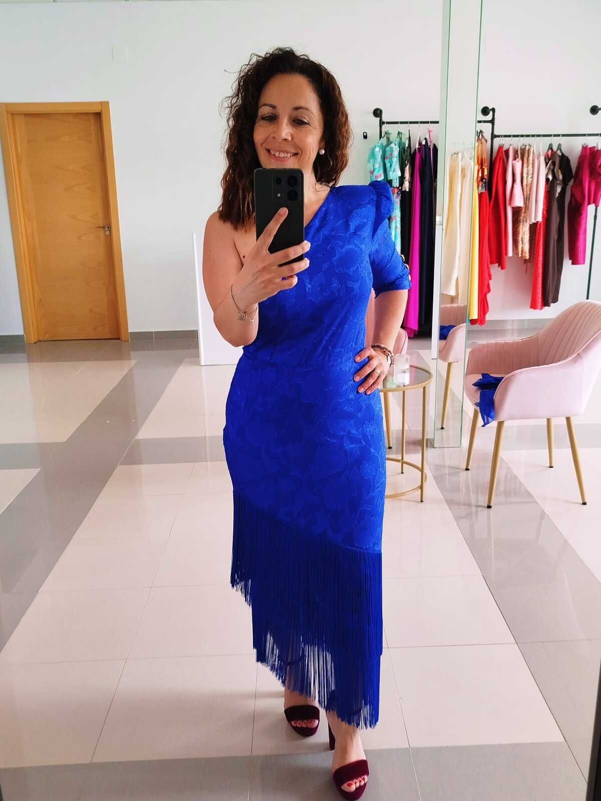 Una mujer con un vestido azul de un solo hombro se toma una selfie frente al espejo. Está en una tienda de ropa con percheros llenos de prendas coloridas.
