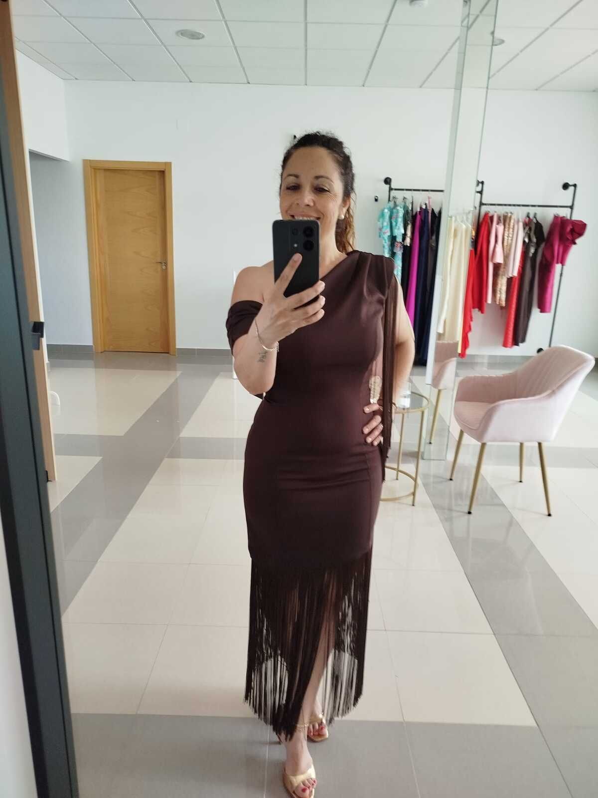 Mujer con un vestido marrón con flecos tomándose una selfie frente al espejo en una tienda de ropa.