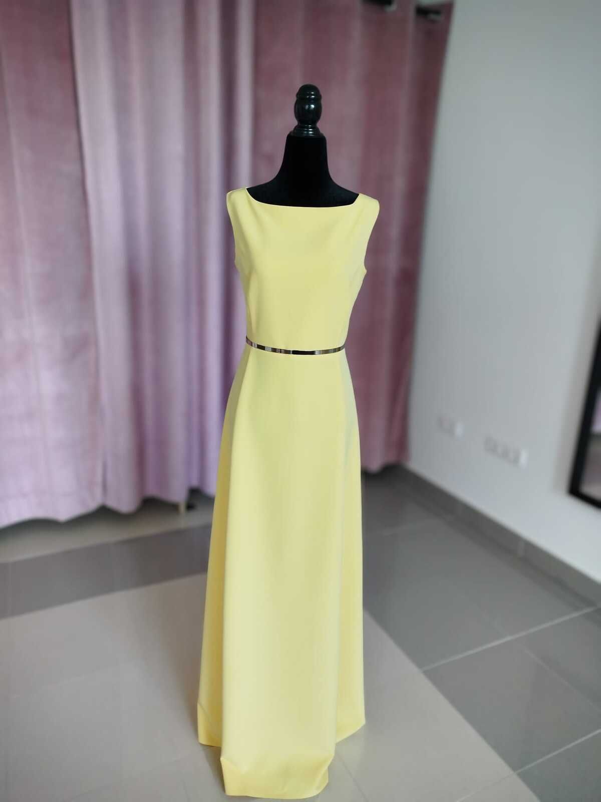 Vestido amarillo largo hasta el suelo con cinturón negro sobre un maniquí negro en una habitación con cortinas rosas.