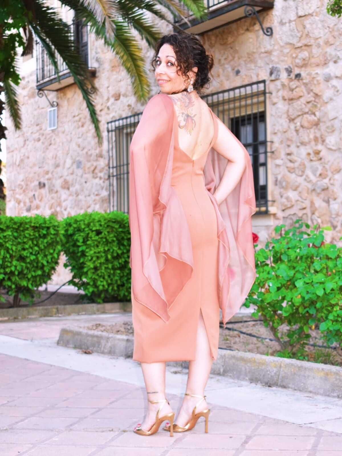 Mujer con vestido color melocotón, tacones y chal posando al aire libre cerca de un edificio de piedra y vegetación.