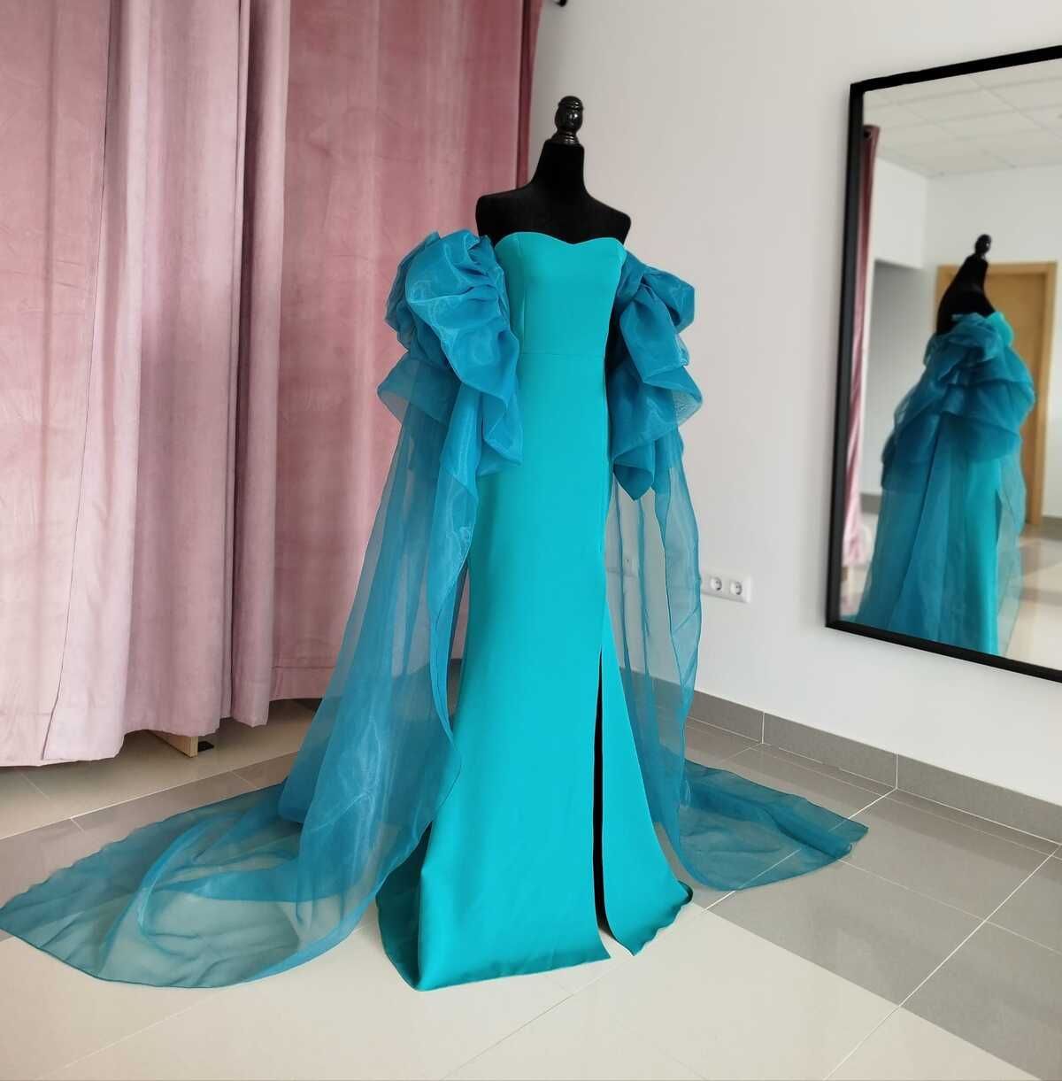 Un vestido turquesa, sin tirantes, con mangas transparentes con volantes y una larga cola sobre un maniquí, en una tienda de ropa.