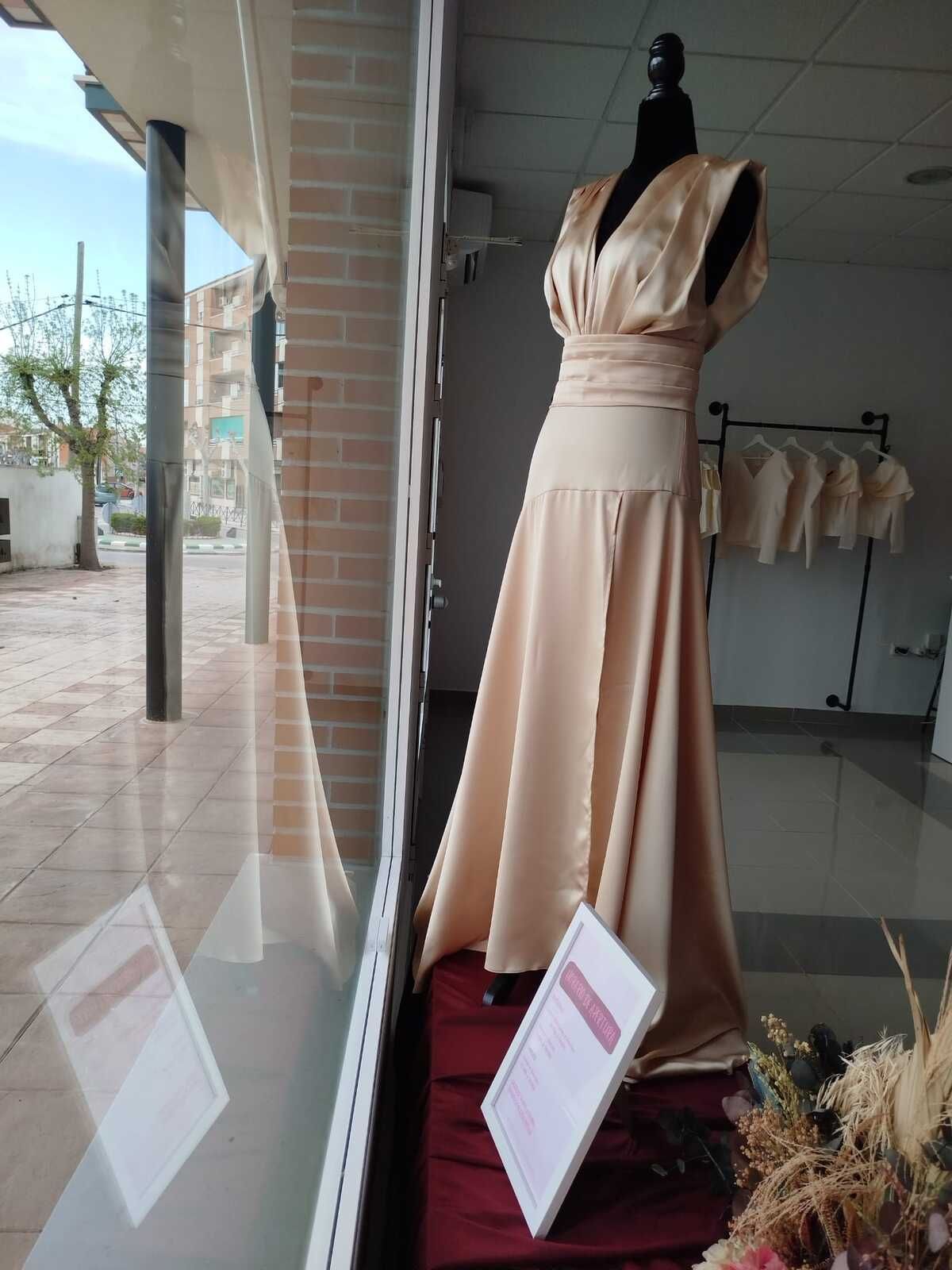 Vestido de satén color canela sobre un maniquí en el escaparate de una tienda, que refleja la escena callejera.