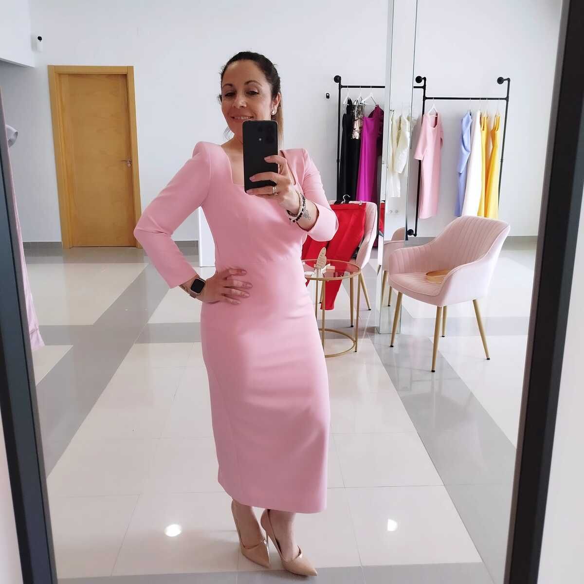 Mujer con vestido rosa tomándose una selfi frente al espejo. De pie en una tienda de ropa, con otras prendas visibles.