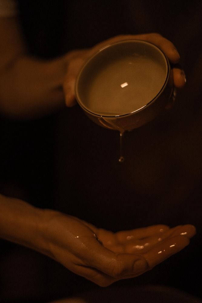 Des mains versent un liquide crémeux d'une tasse dans une paume ouverte, dans une lumière chaude et tamisée.