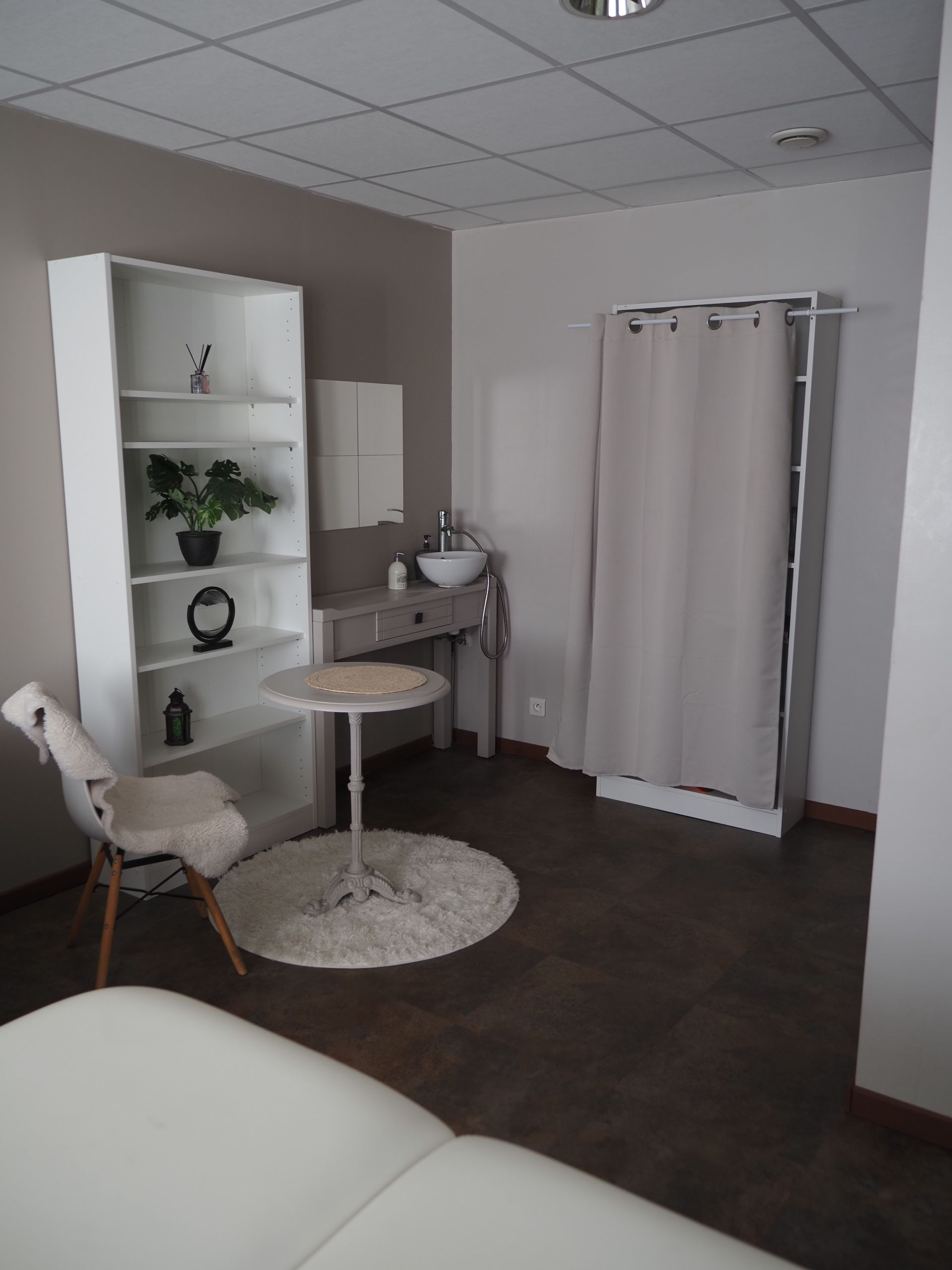 Salle de spa calme avec chaises blanches, petite table, étagères et porte à rideaux sur un sol foncé.