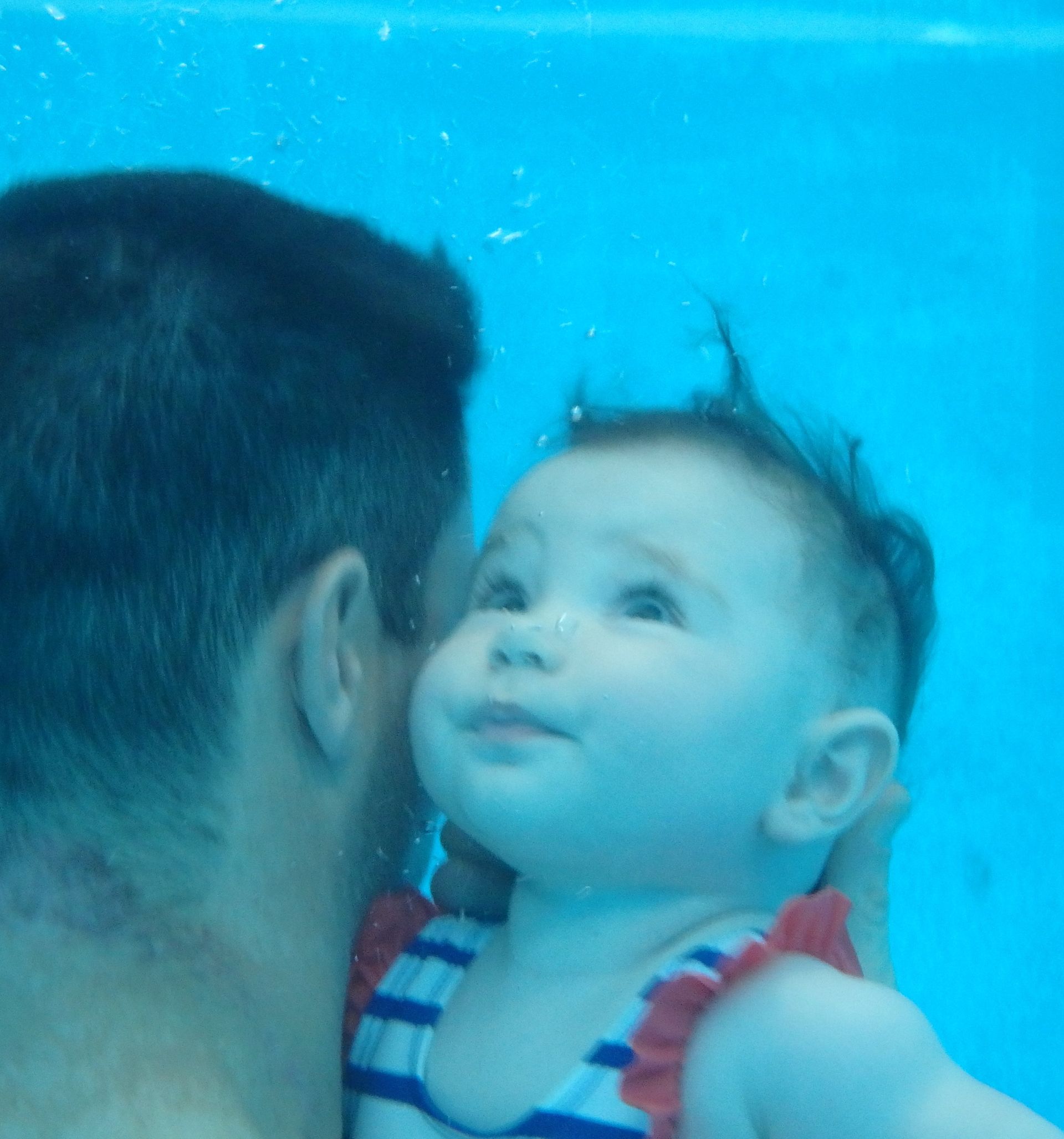 Un homme tenant un bébé dans l'eau bleue, tous deux regardant vers le haut sous l'eau.