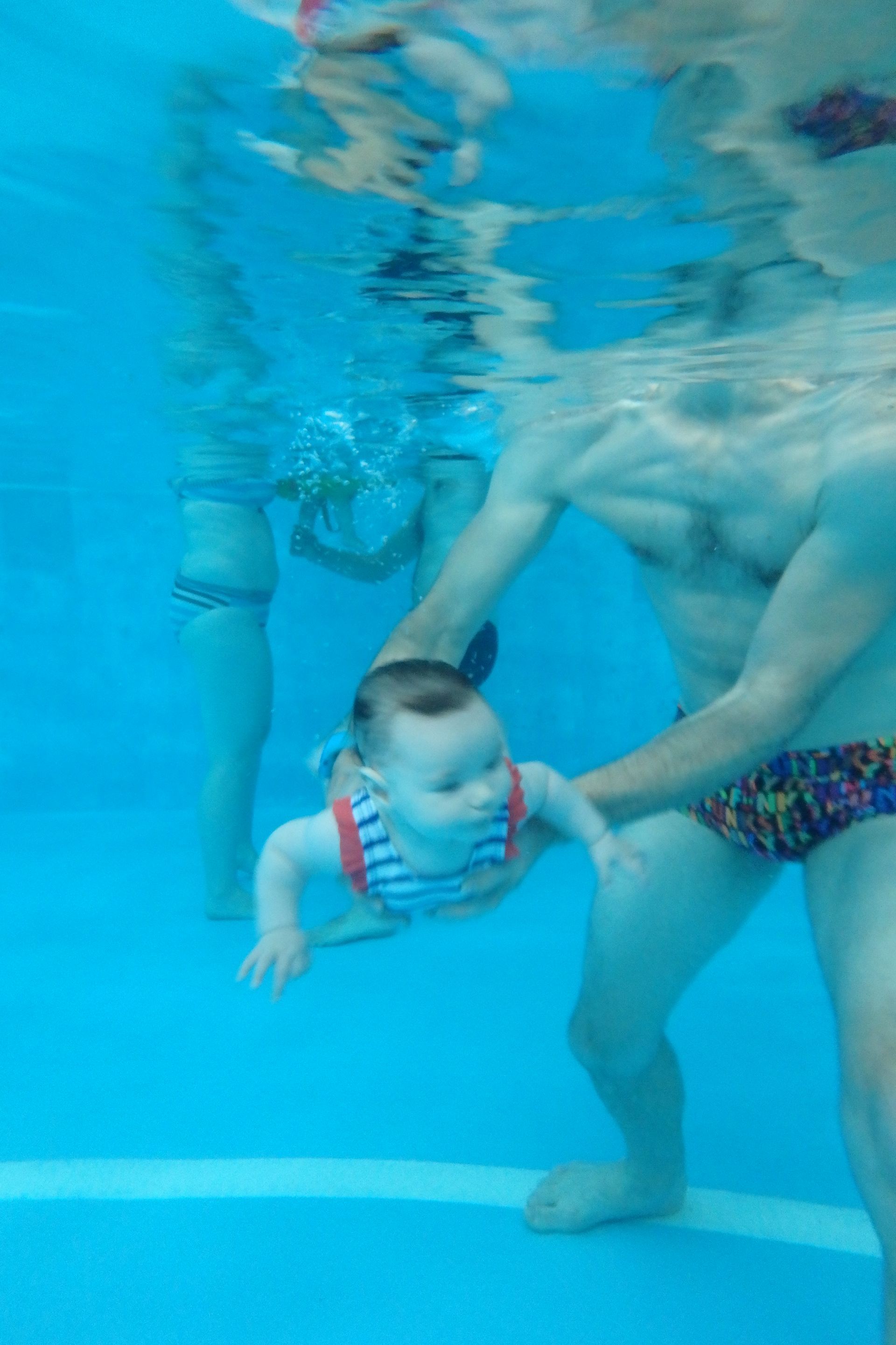 Deux personnes sous l'eau dans une piscine, l'une tenant un bébé face à la caméra.