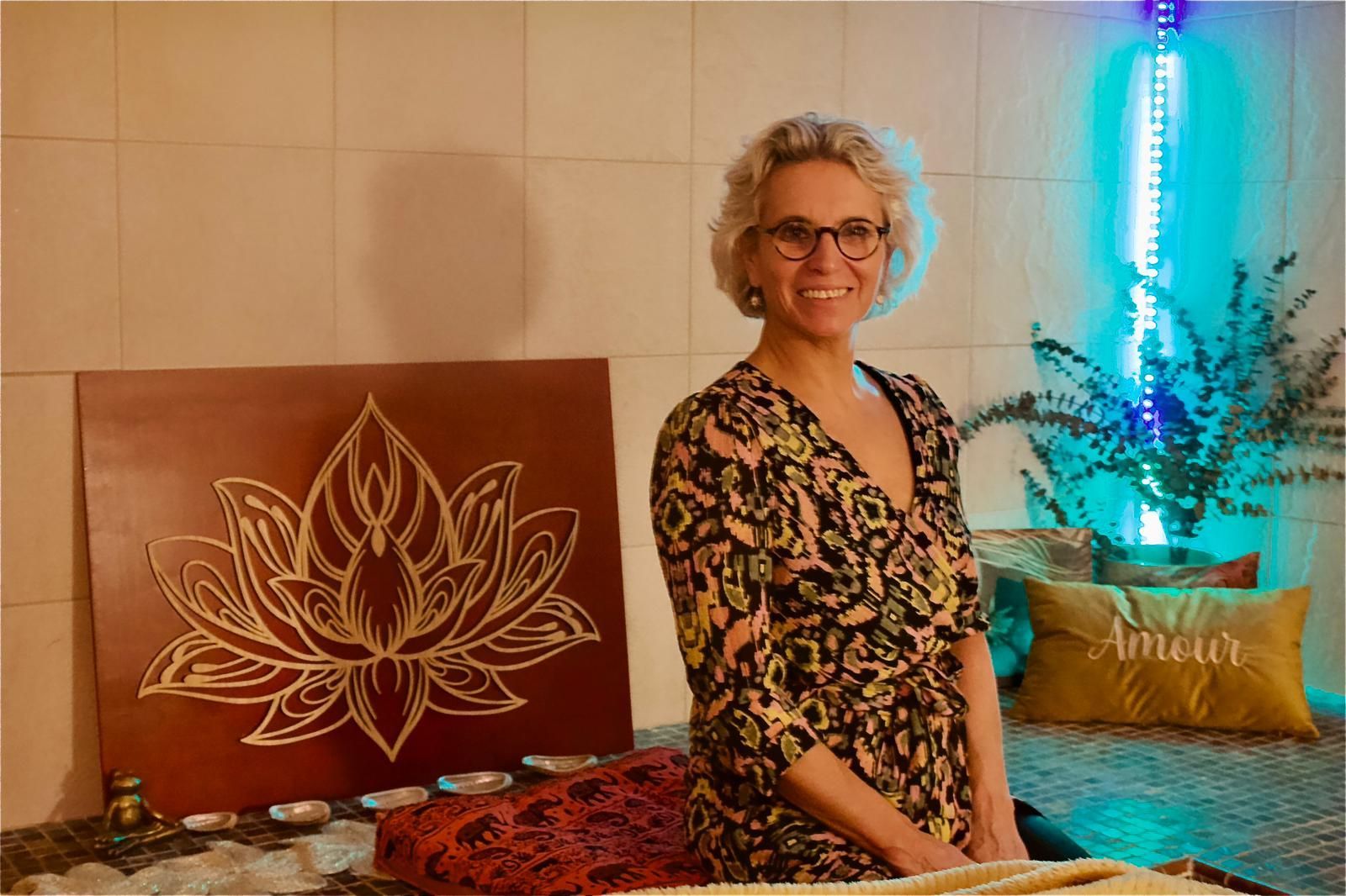 Une personne sourit à côté d'un tapis de yoga orné d'un lotus dans une pièce à la lumière tamisée.