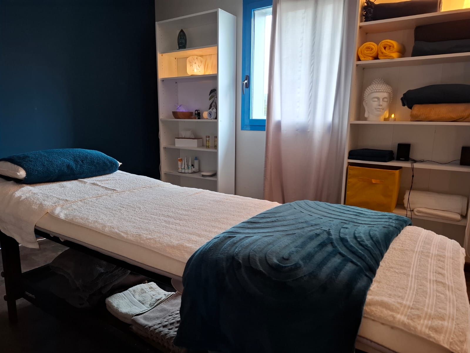 Une table de massage dans une salle de thérapie à l'éclairage tamisé, avec des coussins bleus et des étagères