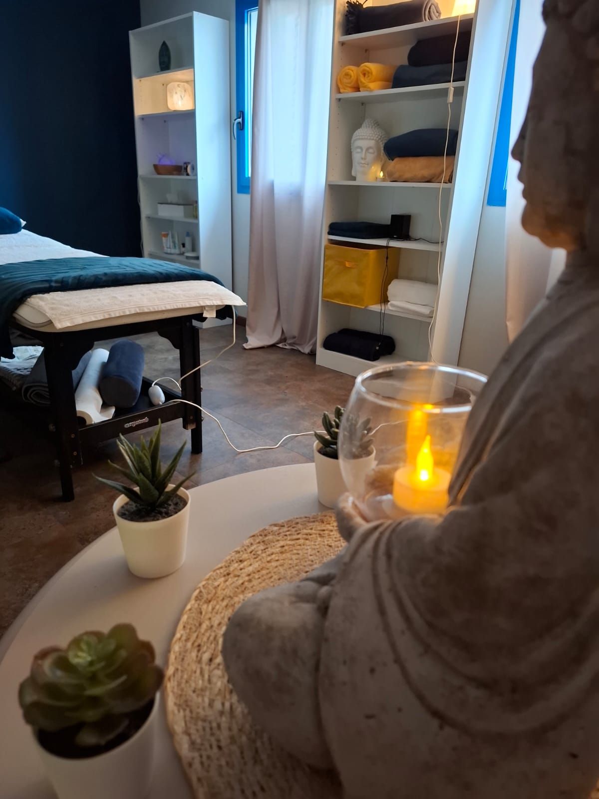 Une personne assise dans une pièce confortable avec une bougie allumée, un lit, des étagères et une petite plante
