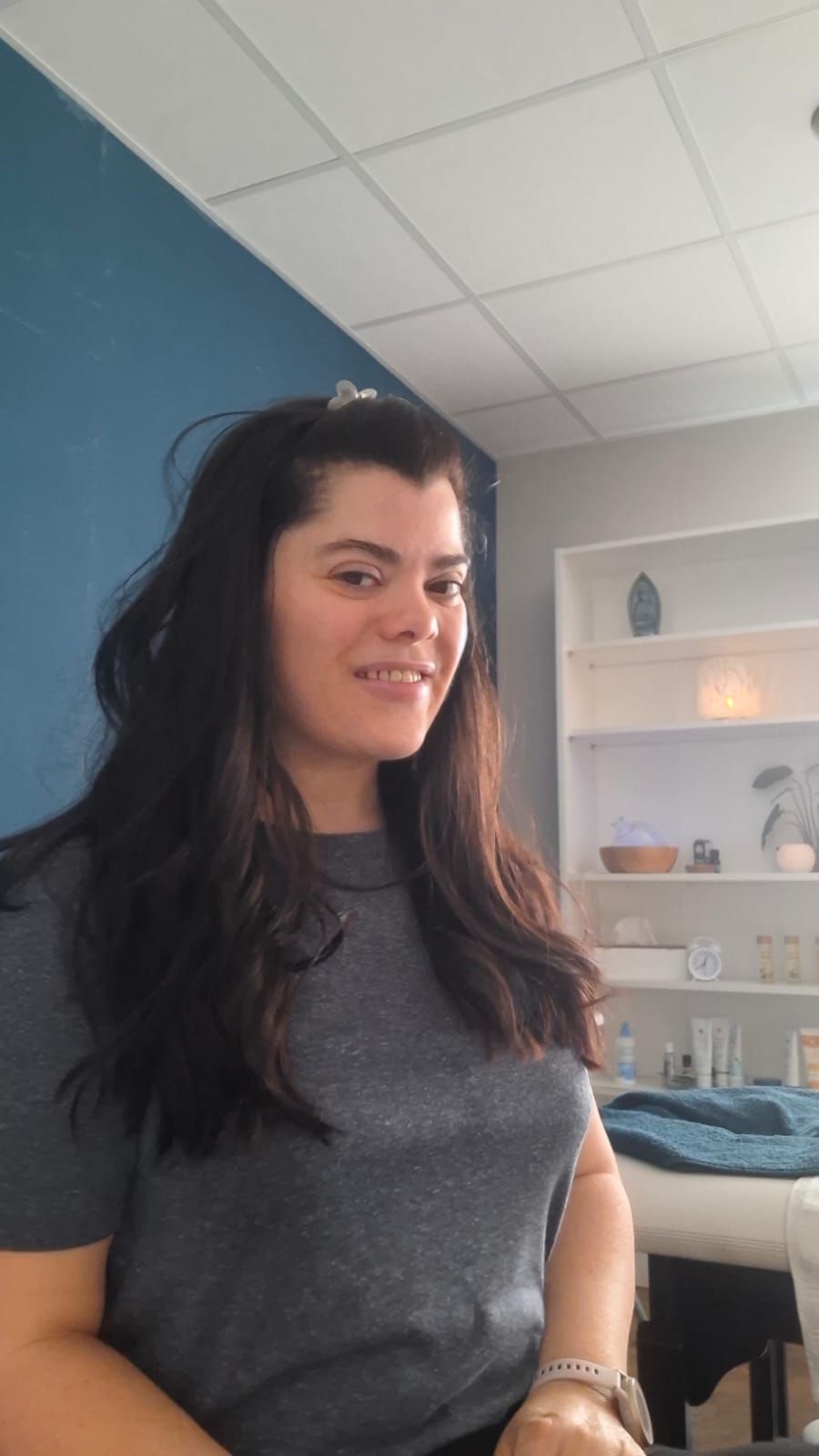 Femme debout dans une pièce lumineuse, souriant légèrement, vêtue d'une chemise gris foncé.