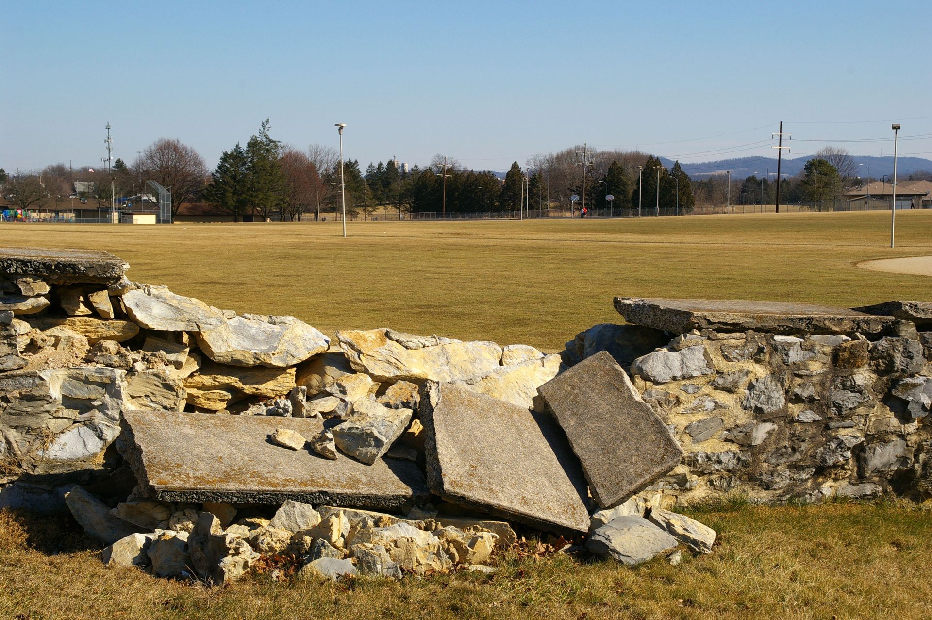 Mur de pierre effondré dans un champ herbeux, avec un bloc de béton brisé au centre et des arbres en arrière-plan.