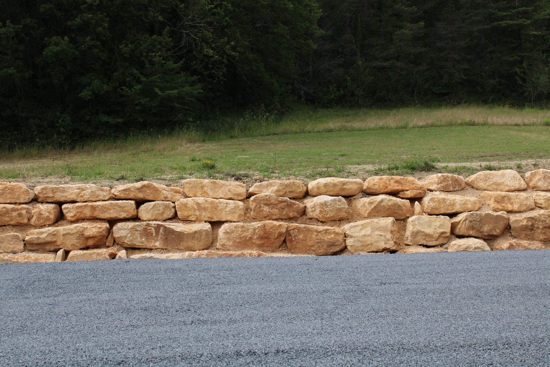 Mur de soutènement en pierre bordant un chemin de gravier, avec un champ herbeux et des arbres en arrière-plan.
