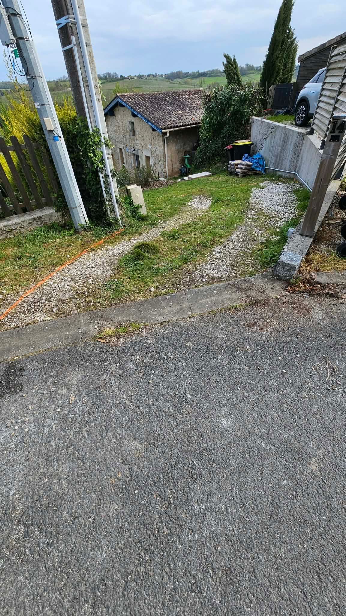 Allée de gravier menant à une petite maison en pierre entourée d'arbres et d'une cour clôturée sous un ciel nuageux.