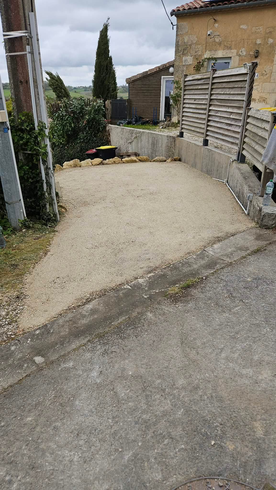 Allée de gravier sinueuse longeant une maison en pierre et une clôture en bois, avec un poteau électrique sur la gauche.