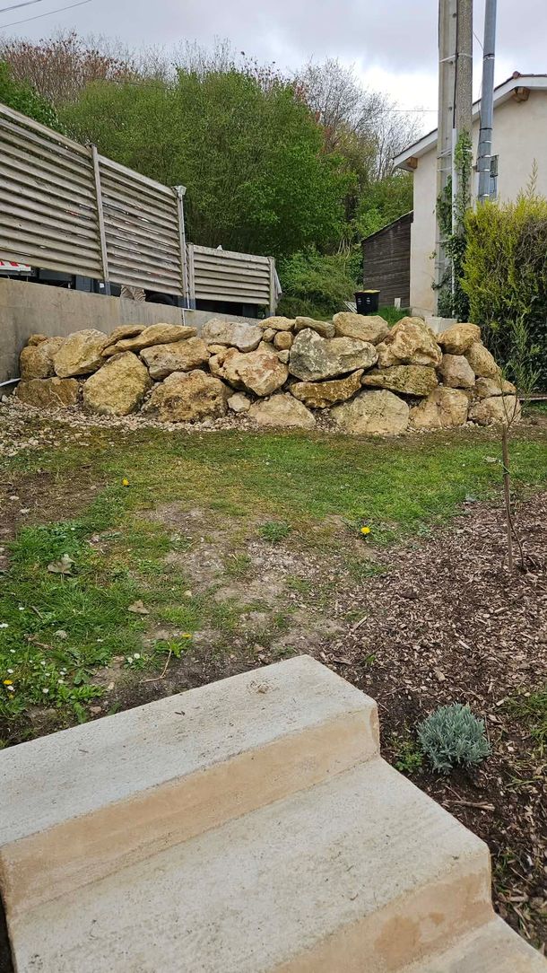 Mur de soutènement en pierre avec allée de jardin en pente, clôture en bois et maison en arrière-plan.