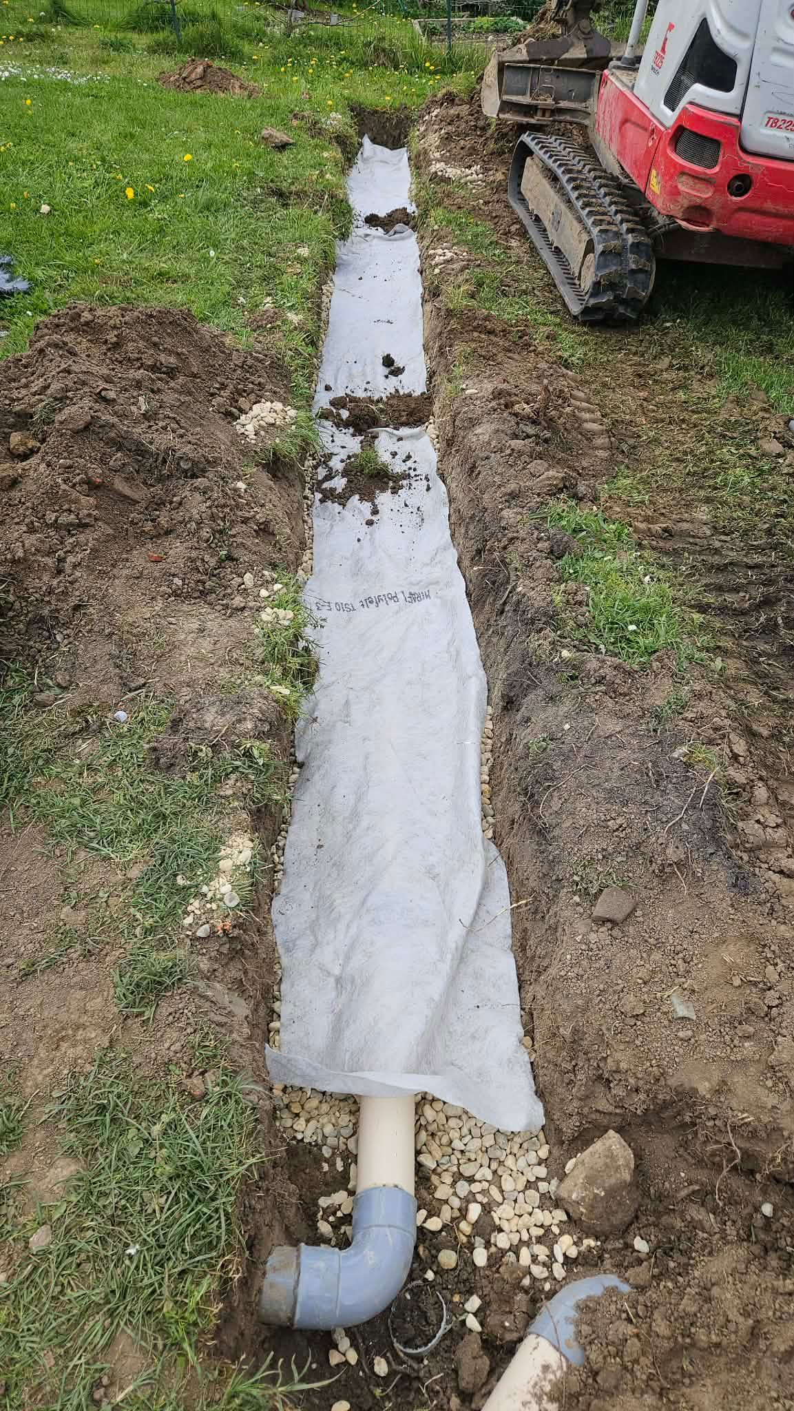 Tranchée de tuyau de drainage enveloppée dans la terre à côté d'une machine de construction rouge.