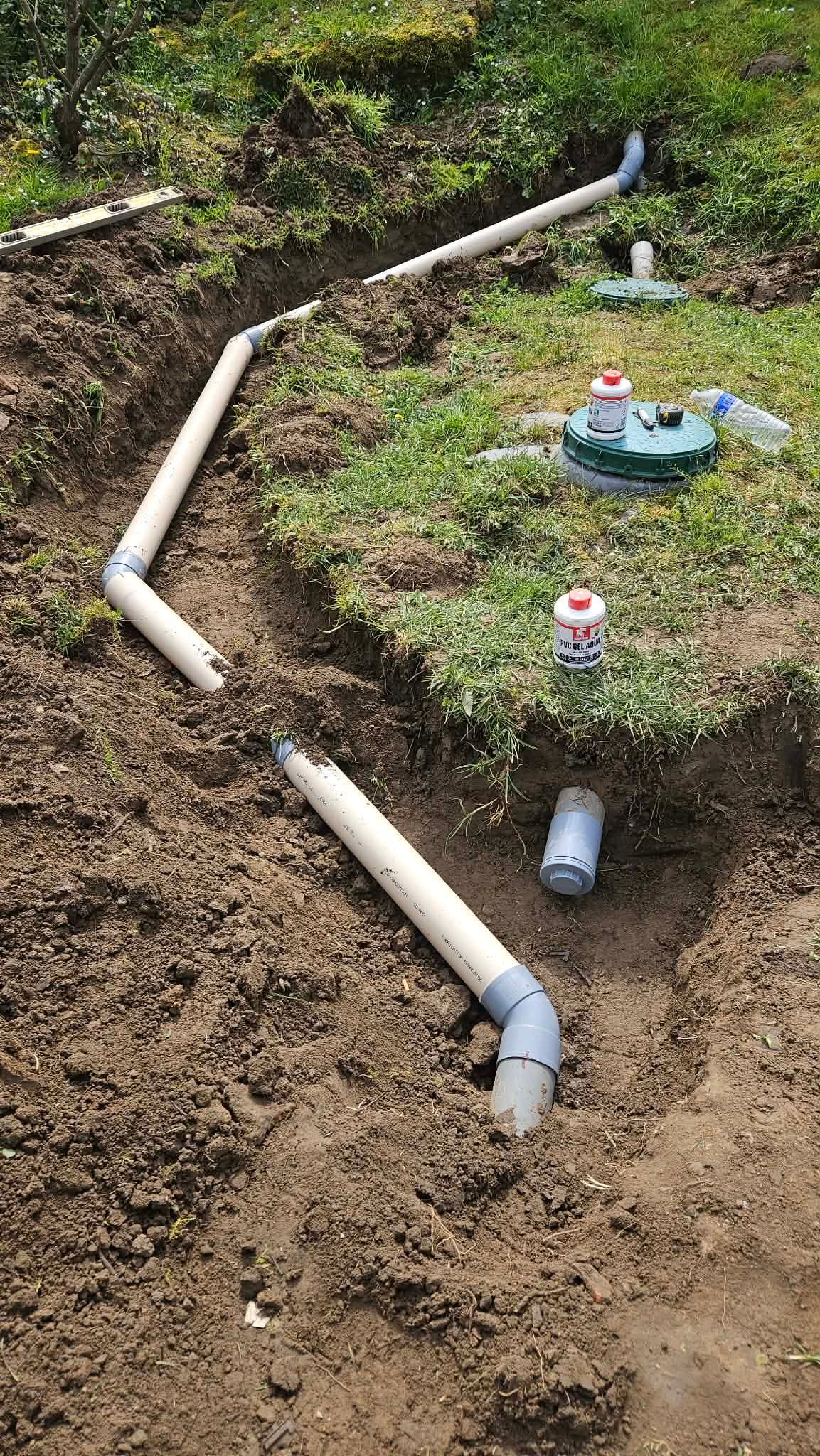 Un tuyau en PVC blanc enterré traverse une tranchée boueuse, à côté de petits arroseurs automatiques et de raccords apparents.