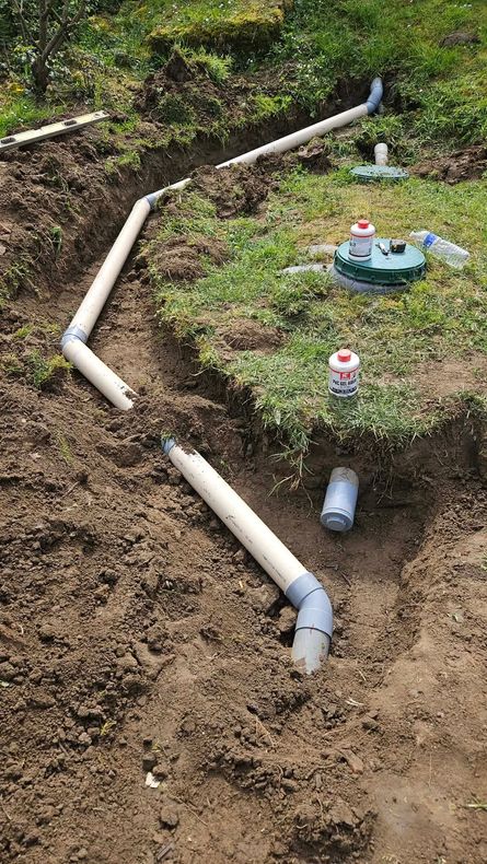 Chemin de terre avec tuyaux en PVC blancs et petits arroseurs dans une cour gazonnée.