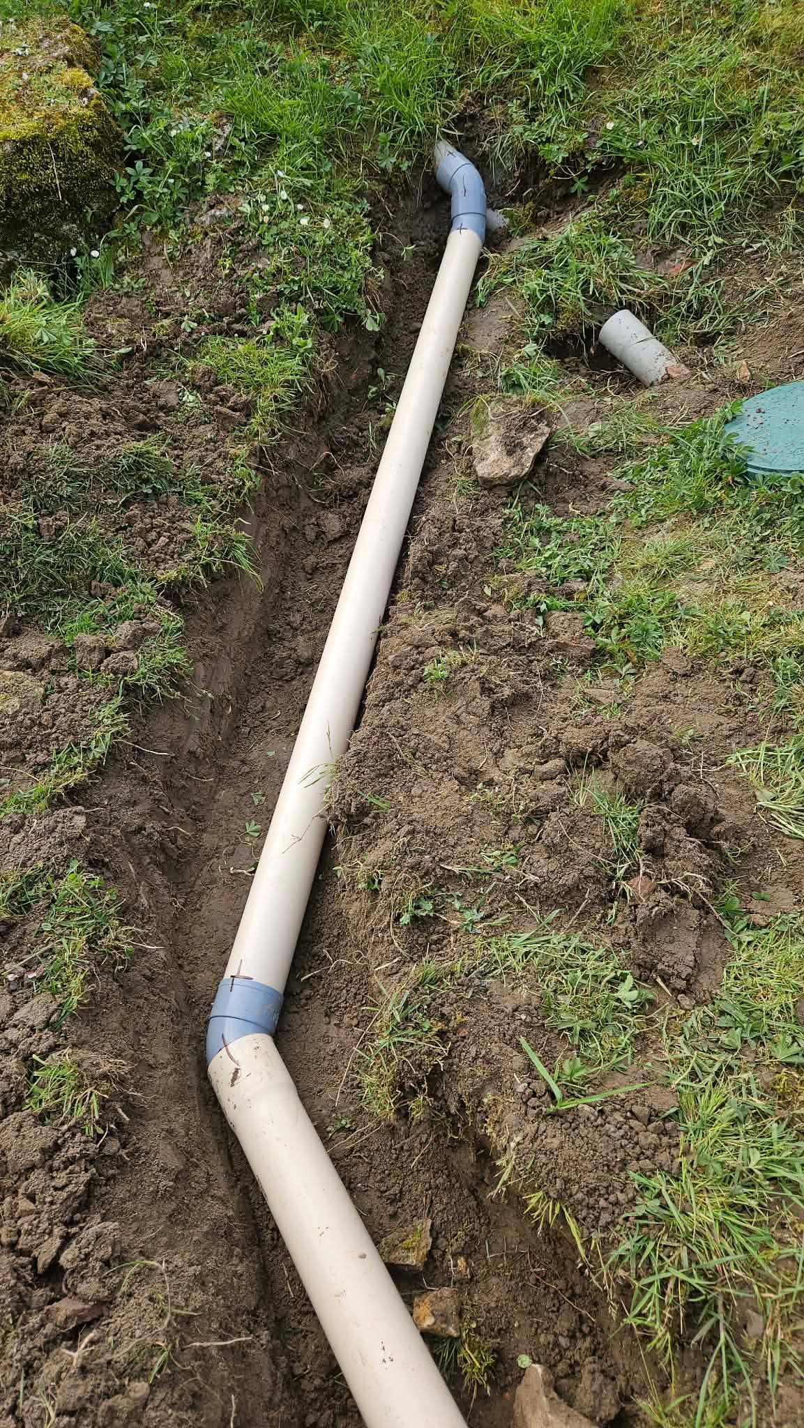 Un morceau de tuyau blanc cassé gît sur l'herbe boueuse, avec un petit morceau de tuyau gris à proximité.