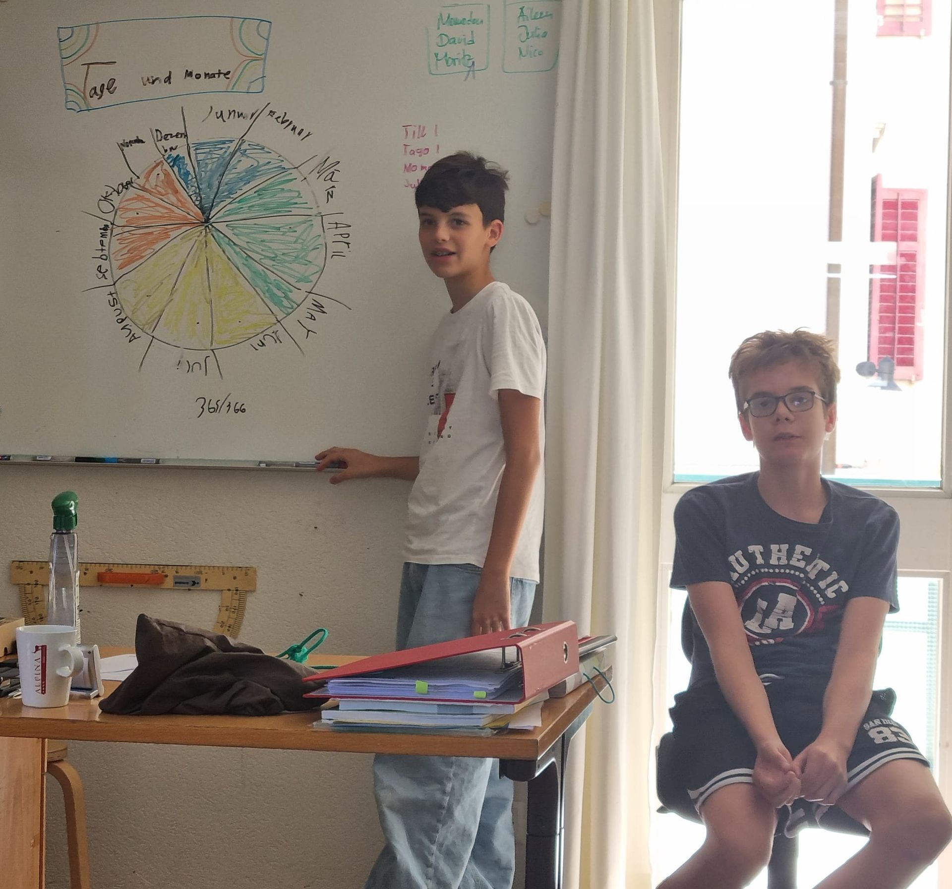 Klassenzimmer mit Tischen, Kunstwerken, Fenstern und Pflanzen. Heller, luftiger Raum in neutralen Farbtönen.