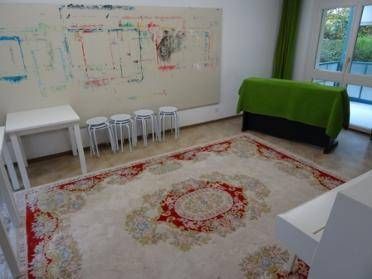 Ein Zimmer mit einem grossen Teppich, Stühlen, Tischen und einem grossen abstrakten Kunstwerk an der Wand.