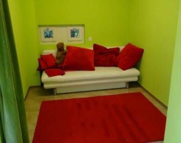 Kleines Zimmer mit weissem Sofa und roten Kissen auf einem roten Teppich vor grünen Wänden.