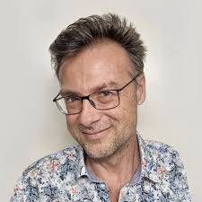 Ein Mann mit Brille lächelt in einem gemusterten Hemd vor einem weissen Hintergrund.