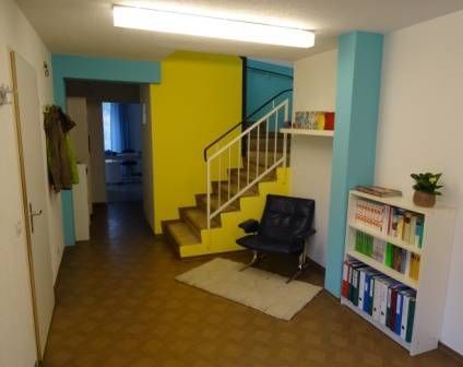 Interieur mit gelben und türkisfarbenen Wänden, Treppe, einem schwarzen Stuhl und einem Bücherregal.