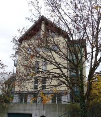 Beiges, mehrstöckiges Gebäude mit braunem Dach hinter einem Baum mit Herbstlaub.