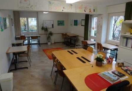 Klassenzimmer mit Tischen, Kunstwerken, Fenstern und Pflanzen. Heller, luftiger Raum in neutralen Farbtönen.