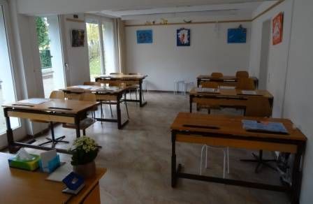 Leeres Klassenzimmer mit Tischen, Stühlen und natürlichem Licht durch grosse Fenster.