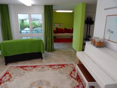 Ein Zimmer mit einem weißen Klavier, einem grünen Tisch und einem bunten Teppich. In einem anderen Bereich steht eine Couch.