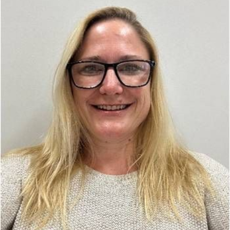 Frau mit blonden Haaren, Brille, lächelnd und in einem grauen Pullover.