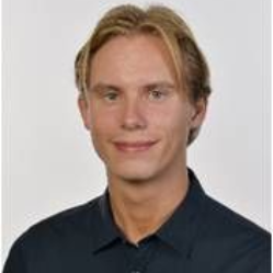 Mann mit blonden Haaren, der ein dunkelblaues Hemd mit Knöpfen trägt, lächelt.