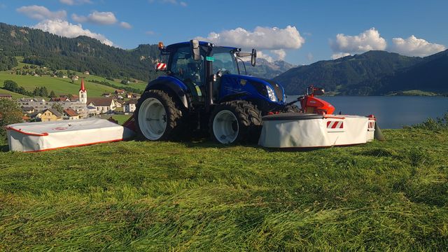 Ein blauer Traktor mäht unter klarem, blauem Himmel einen grünen Grashang.