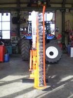 Eine orange-gelbe Landmaschine mit langem Schneidmesser steht vor einem blauen Traktor in einer Werkstatt.