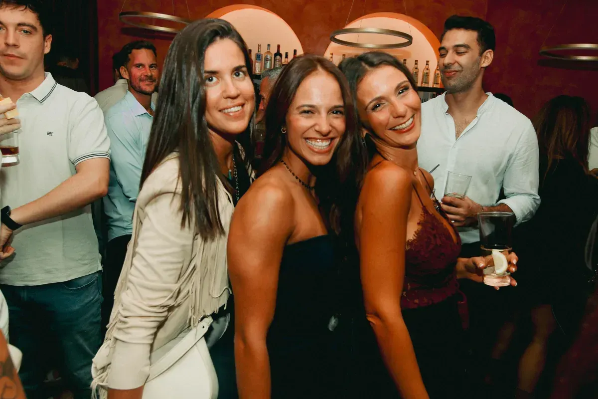 Tres mujeres sonrientes posan en un bar con otras personas al fondo.