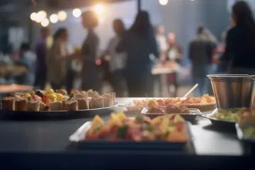 Mesa de bufé con aperitivos en una reunión al aire libre; personas borrosas al fondo.